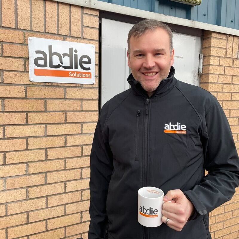 A scottish engineer in a black jacket holding an Abdie Solutions mug